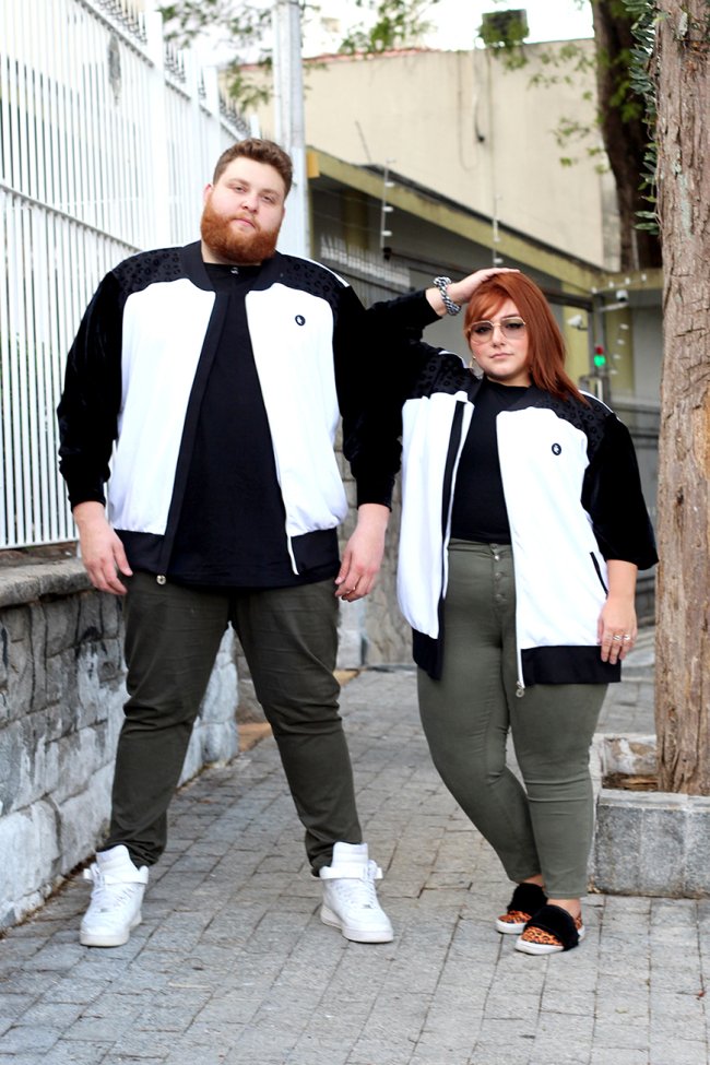 Casal posando na rua com blusa de frio preto e branca igual, camiseta preta igual, e calça verde militar igual. Os dois estão com aparência séria.