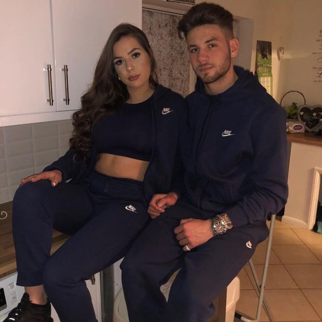 look combinando preto Casal sentado em bancos na cozinha, usando roupas parecidas. A menina usa top preto, blusa de moletom e calça preta. O menino usa blusa e calça de moletom. Os dois estão com uma expressão séria.