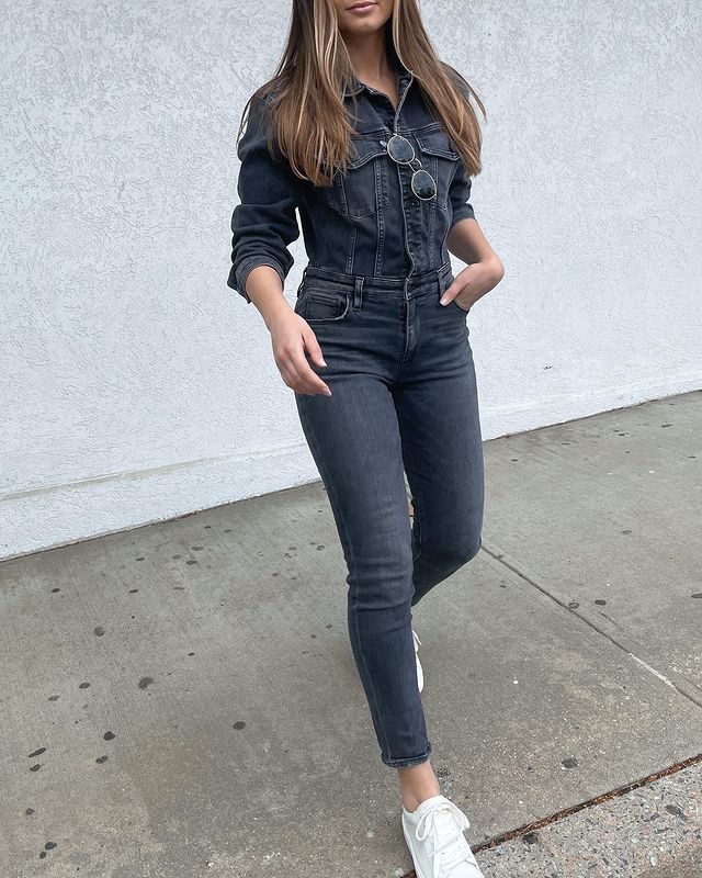 macacao-jeans-cristielaratta Garota usando macacão jeans em lavagem escura com tênis branco e um óculos de sol pendurado na parte de cima da roupa. A foto está cortada na altura da boca e ela está andando em direção à câmera.