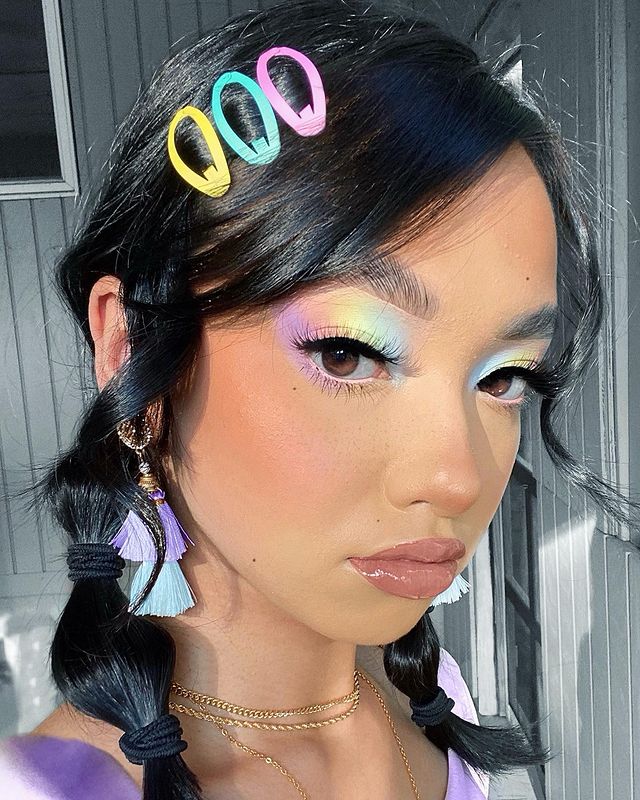 make em tons pastel Foto de uma mulher. Ela usa o cabelo em um penteado bolha, com presilhas coloridas de cabelo, maquiagem com gloss, pele natural e sombra com mix de cores em tons pastel. Ela olha para a câmera e não sorri.