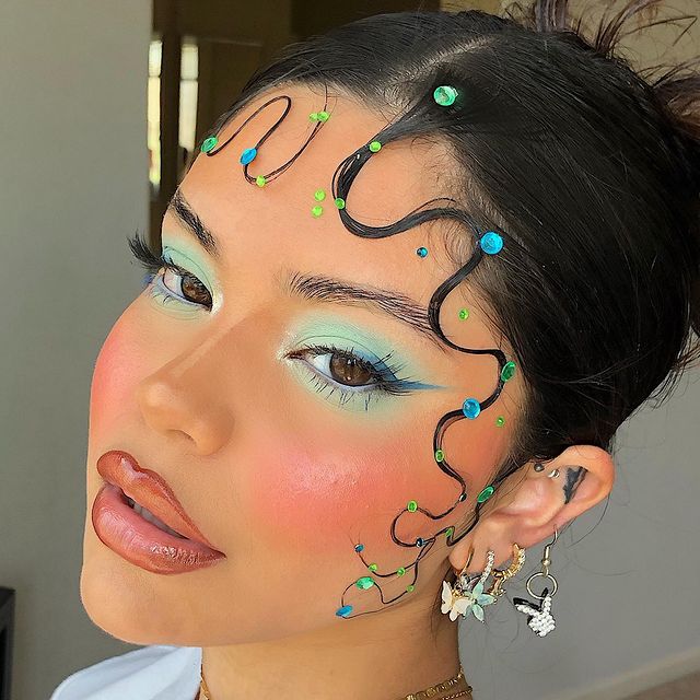 maquiagem azul pastel Selfie de uma mulher com o rosto de perfil. Ela usa uma camiseta azul, cabelo modelado em um baby hair com uma decoração azul e verde, maquiagem com sombra e delineado azul. Ela olha para a câmera e não sorri.