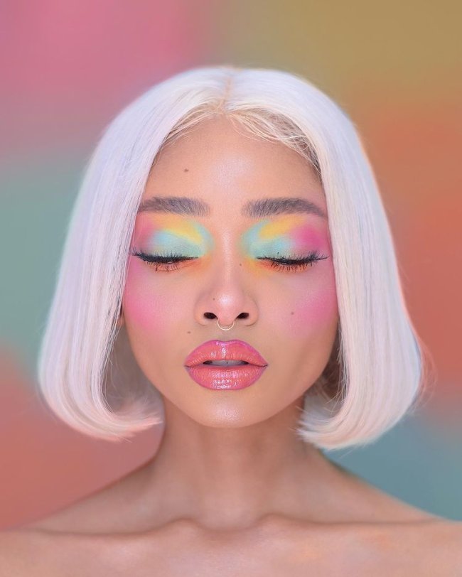 mix de sombra em tons pastel Foto de uma mulher. Ela usa o cabelo curto solto, maquiagem com gloss, blush rosa marcado e sombra em tons de verde, rosa e amarelo pastel. Ela está de frente para a câmera, de olhos fechados e não sorri.