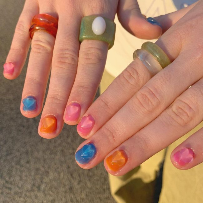 Foto com foco nas mãos, com esmalte colorido. Unhas laranja, rosa, azul e rosa. Com coração em 3D.