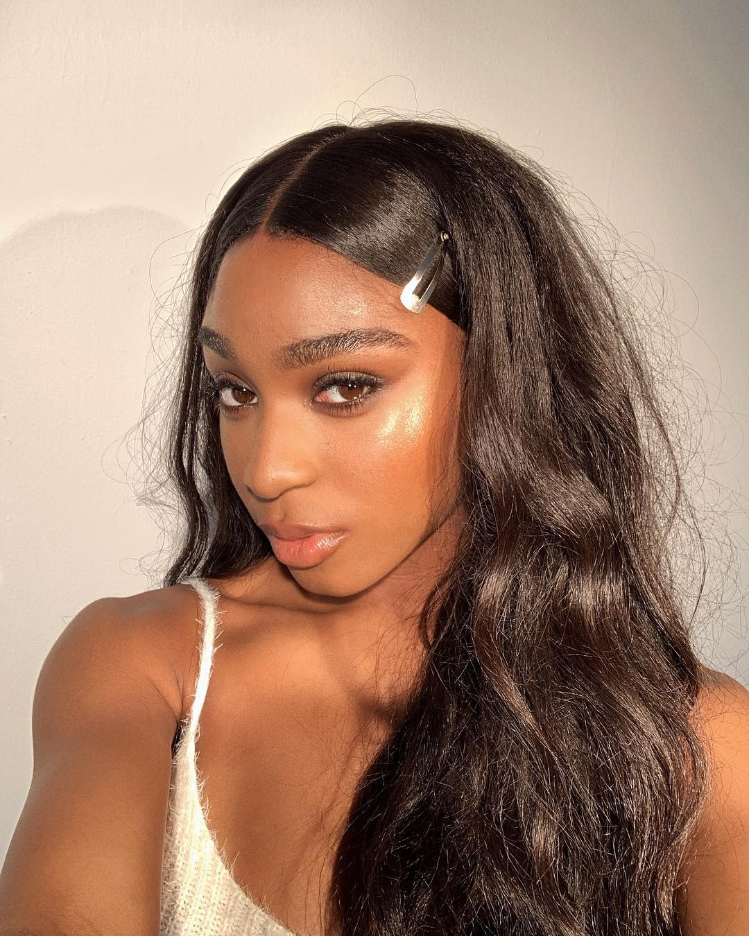 Selfie de Normani posando com expressão séria; ela usa uma regata branca com maquiagem iluminada e uma presilha prata prende parte de sua franja