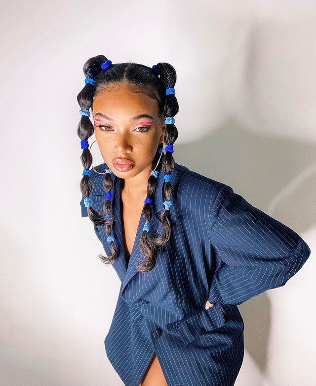 Foto de uma mulher em frente a uma parede branca. Ela usa um blazer azul com riscos brancos, cabelo preso em um penteado bolha dividido em quatro partes. Ela está com as mãos na cintura, olha para a câmera e não sorri.