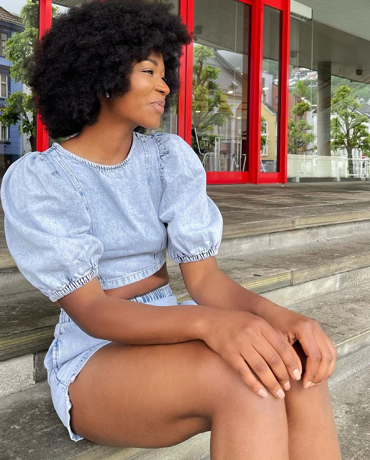top-blusa-jeans-mabel.sama Garota usando top cropped jeans e short jeans. Ela está sentada em uma escada, com as duas mãos apoiadas nos joelhos, olhando para o lado e dando um leve sorriso sem mostrar os dentes.