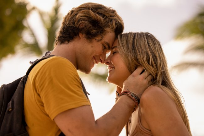 John B. e Sarah em cena de Outer Banks; eles estão sorrindo com as testas e os narizes encostados; John B. está com as mãos no pescoço da personagem