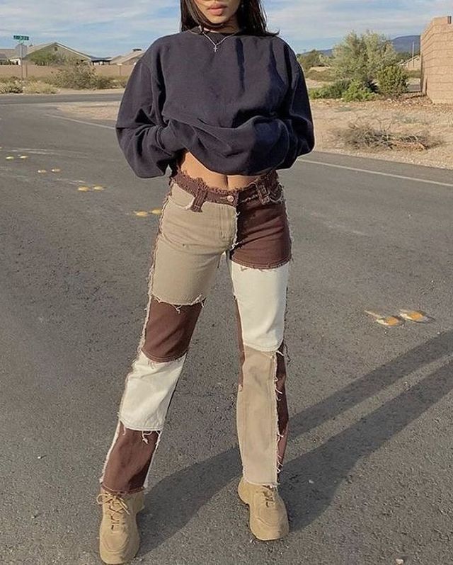 Patchwork marrom Na imagem não conseguimos ver o rosto de quem usa as peças, somente a blusa de moletom cinza escuro e a calça com detalhes em patchwork. A pessoa posa no meio da rua