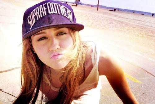 bone-de-aba-reta Foto da cantora Miley Cyrus. Ela usa um boné de aba reta e camiseta branca. Ela faz pose com um bico para a foto.