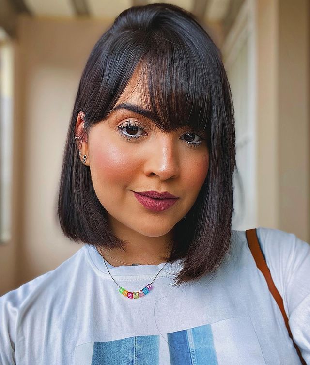 Jovem sorridente com camiseta branca posando para selfie. Ela tem cabelo chanel preto com franja na altura dos olhos.