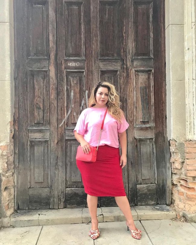 Jovem posando em frente a porta de madeira, usando camisa rosa e saia vermelha e uma bolsa transpassada.