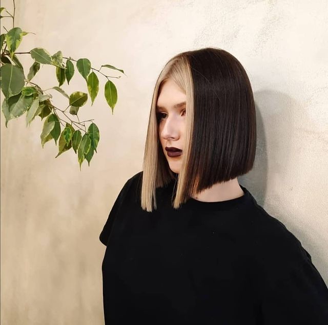 Jovem posando com roupa preta, perto a folhas verde, com parede bege de fundo e expressão séria. Seu cabelo tem as mechas da frente platinadas e as detrás são pretas.