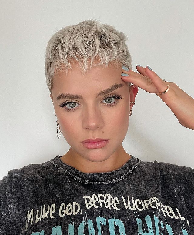 corte pixie cabelo platinado Jovem de camiseta cinza, posando para foto com expressão séria e uma das mãos apoiada no rosto.