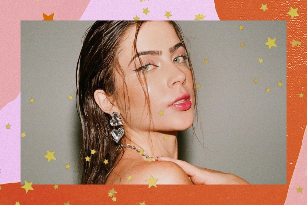 Montagem com a foto da influenciadora Jade Picon com o fundo rosa e laranja com detalhes de estrelas douradas. Ela usa o cabelo escuro solto, colar, brinco e olha para a câmera sem sorrir.