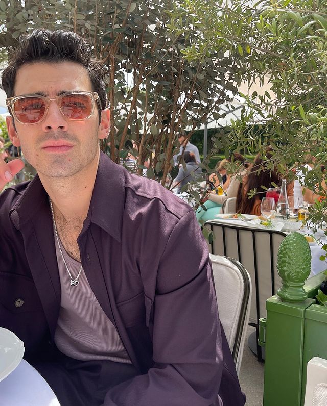 Joe Jonas olhando para algo atrás da câmera; ele usa uma camiseta lilás com uma camisa roxa por cima e óculos de sol com lente vermelha; sua expressão é confusa