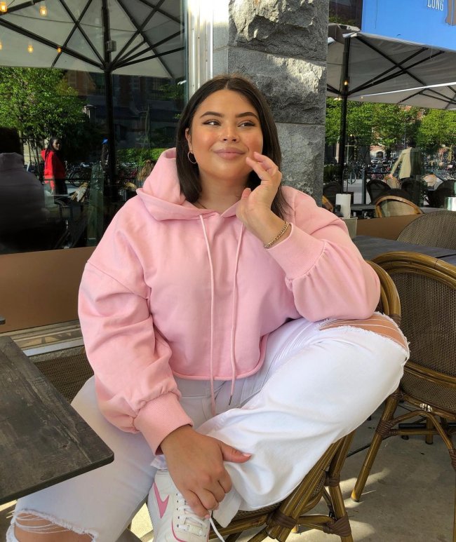 Foto de uma mulher sentada em uma mesa de restaurante. Ela usa uma blusa de moletom com capuz rosa, calça jeans branca e tênis branco. Ela apoia a mão esquerda no queixo e a segura a perna esquerda com a mão direita, olha para o lado e sorri levemente.