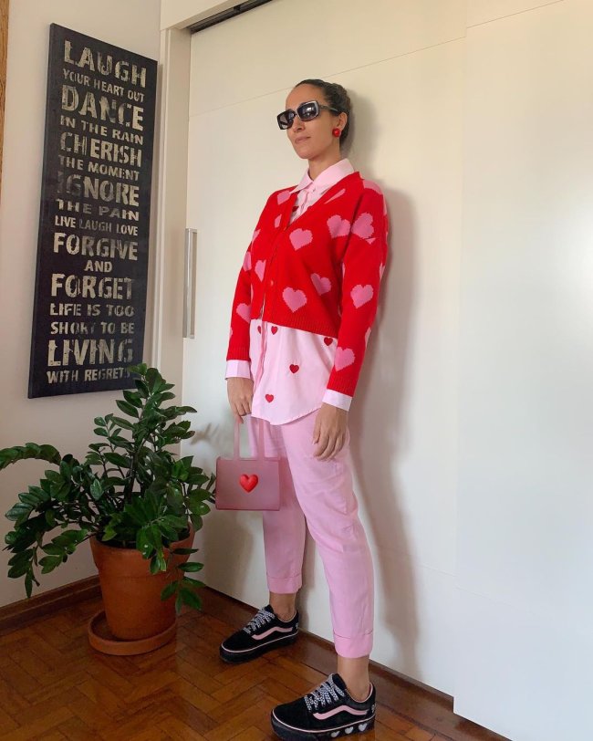 look rosa e vermelhp Jovem encostada na parede usando casaco vermelho, camisa rosa e calça rosa. Sua expressão é séria. Podemos ver um vaso de planta ao lado dela e um quadro preto com frases escritas na cor branca.