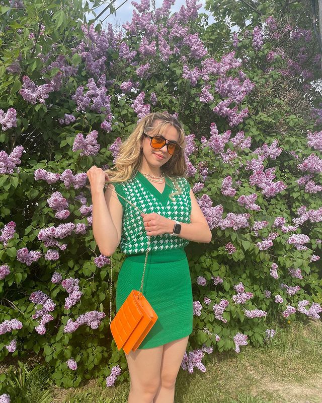 Garota usando colete de tricô sem alças verde e saia no mesmo tom, além de um óculos de sol de lentes alaranjadas e uma bolsa laranja neon, que está sendo segurada pelas suas duas mãos. Ao fundo dela, há várias flores roxas.