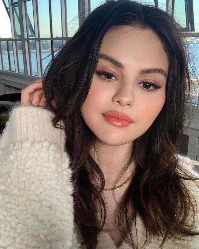 Selfie da atriz e cantora Selena Gomez. Ela usa um casaco bege de pelinhos, cabelo solto, olho esfumado, gloss e uma corrente dourada. Ela olha para a câmera, sorri levemente e está com a mão direita perto do rosto.