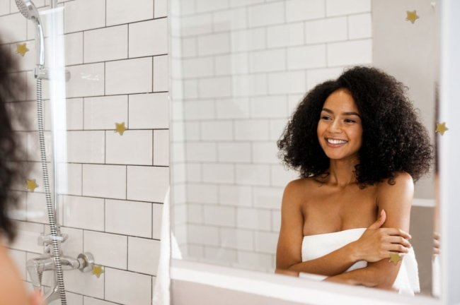 skincare Jovem sorridente enrolada em toalha se olhando no espelho. Ela está dentro de um banheiro branco.