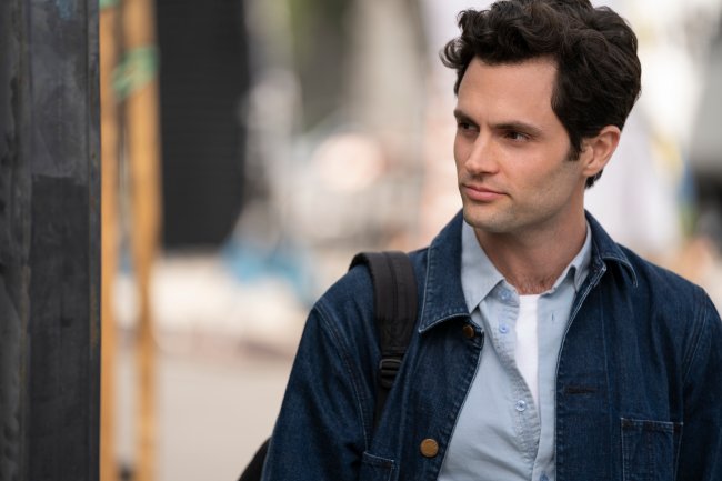 Love e Joe na segunda temporada de Você (You), série da Netflix; eles estão olhando para frente com expressões sérias, um do lado do outro; Joe está com as costas viradas para Love, que usa uma blusa vermelha com cabelo solto; Joe usa uma camiseta branca com uma camisa clara por cima