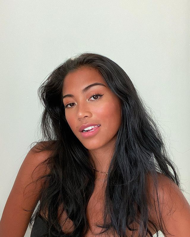cabelo-castanho-escuro Selfie de uma mulher. Ela usa um top cropped preto, colarzinho dourado, está com o cabelo solto e maquiagem natural. Ela olha para a câmera e sorri levemente.