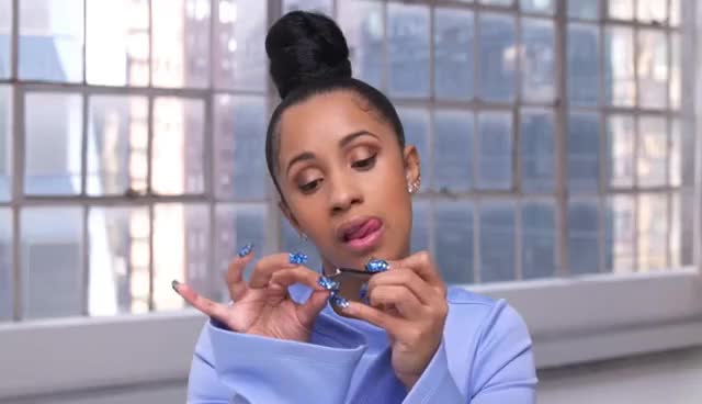 Cardi B usando blusa azul de manga comprida, um coque no topo da cabeça, olhando para baixo enquanto faz as unhas.