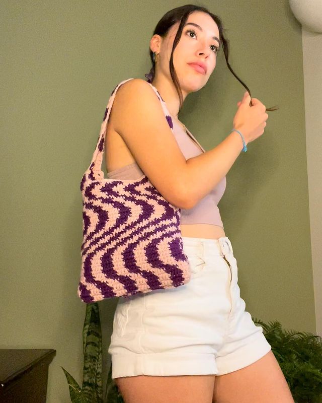 ecobag-de-croche Foto de uma mulher em uma sala. Ela usa um top cropped lilás, short jeans branco, ecobag lilás e roxa de crochê e cabelo preso em um coque. Ela segura um fio da franja com a mão direita, olha para frente e não sorri.