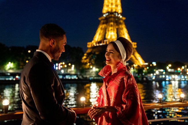 emily em paris Imagem da segunda temporada de Emily em Paris, série da Netflix; Alfie e Emily estão conversando e sorrindo um para o outro; ele usa um terno preto e ela usa um casaco rosa com o cabelo preso com uma faixa branca; o fundo mostra o rio e as luzes da Torre Eiffel