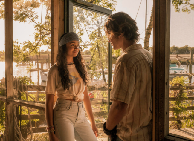 Kiara conversando com John B. Ela esta de frente e sorridente e ele de costas. Ela usa faixa na cabeça e camiseta.