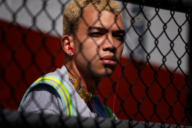 EXT HIGH SCHOOL – TRACK FIELD – EP 101 Personagem de Justice Smith observando algo a sua frente através de uma grade em formato de losangos