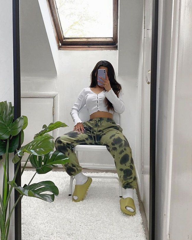 Jovem posando sentada com calça camuflada verde e blusinha branca. O celular cobre seu rosto.