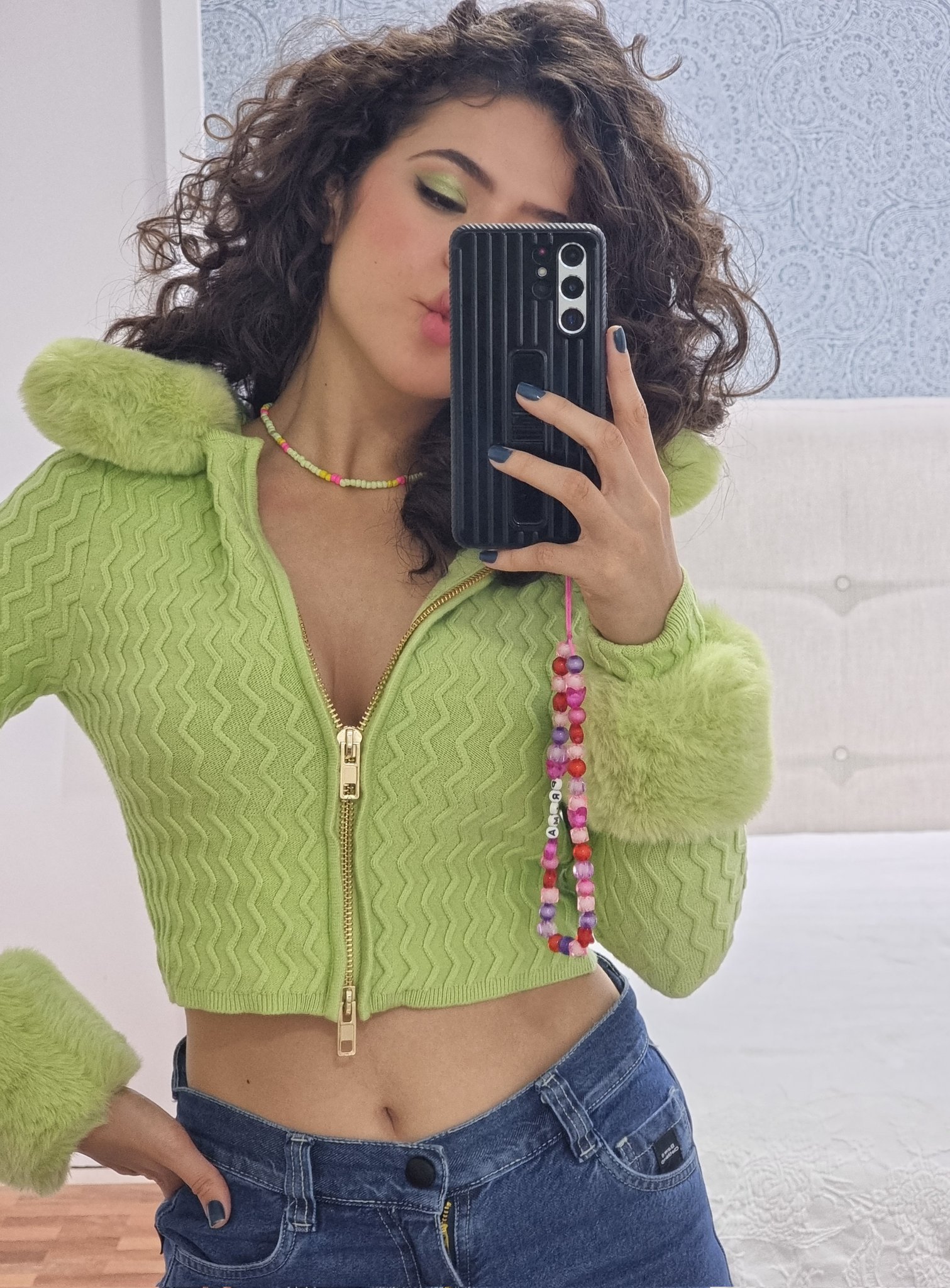 Maisa Silva usando cardigan verde cropped e calça jeans com celular na frente do rosto em frente ao espelho