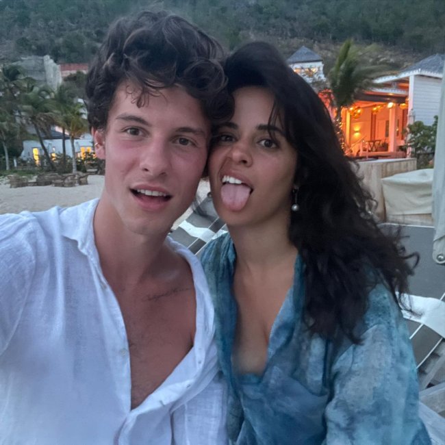 Shawn Mendes e Camila Cabello em selfie na praia tirada pelo cantor; os dois estão com os cabelos bagunçados e fazendo careta; Shawn usa uma camisa branca aberta e Camila está com uma saída de praia azul com detalhes brancos mostrando a língua