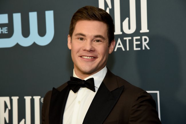 Adam Devine no Critics' Choice Awards em 2020; ele usa um terno preto com gravata borboleta preta e camisa branca; ele está posando e sorrindo para a câmera no tapete vermelho da premiação; a imagem mostra apenas do peito para cima do ator