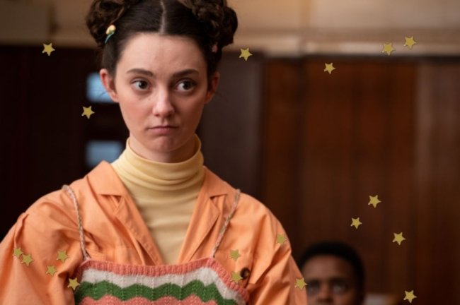 SPACE BUNS Lily, de Sex Education, com space buns, ela usa blusa bege e camisa laranja.