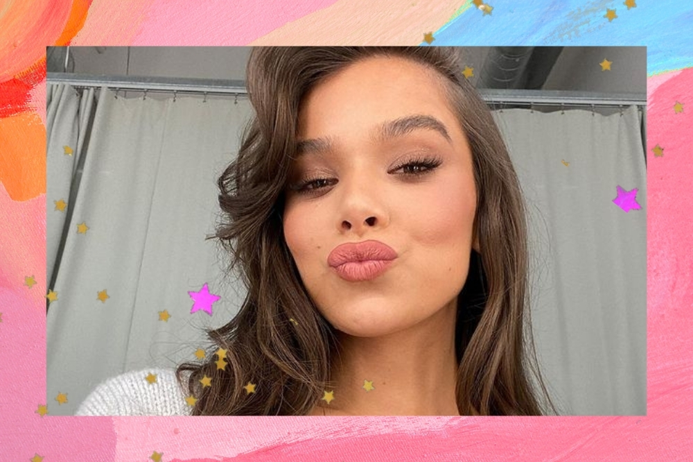 Montagem com o fundo rosa, laranja e azul com detalhe de estrelas douradas com uma selfie da cantora e atriz Hailee Steinfeld. Ela está com o cabelo solto, maquiagem básica, olha para a câmera e faz bico.