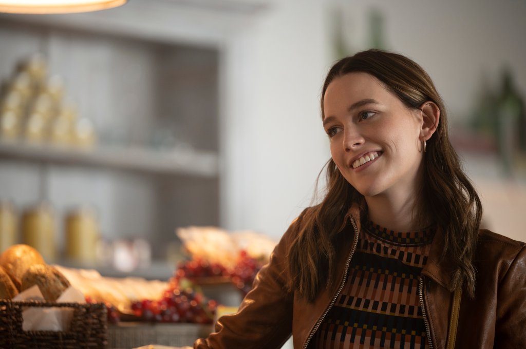 Victoria Pedretti como Love Quinn na série Você; ela está sorrindo e olhando para o lado com uma jaqueta marrom e uma blusa de tricô com listras horizontais em tons de marrom e preto; ao fundo é possível ver alguns pães e doces da padaria da personagem