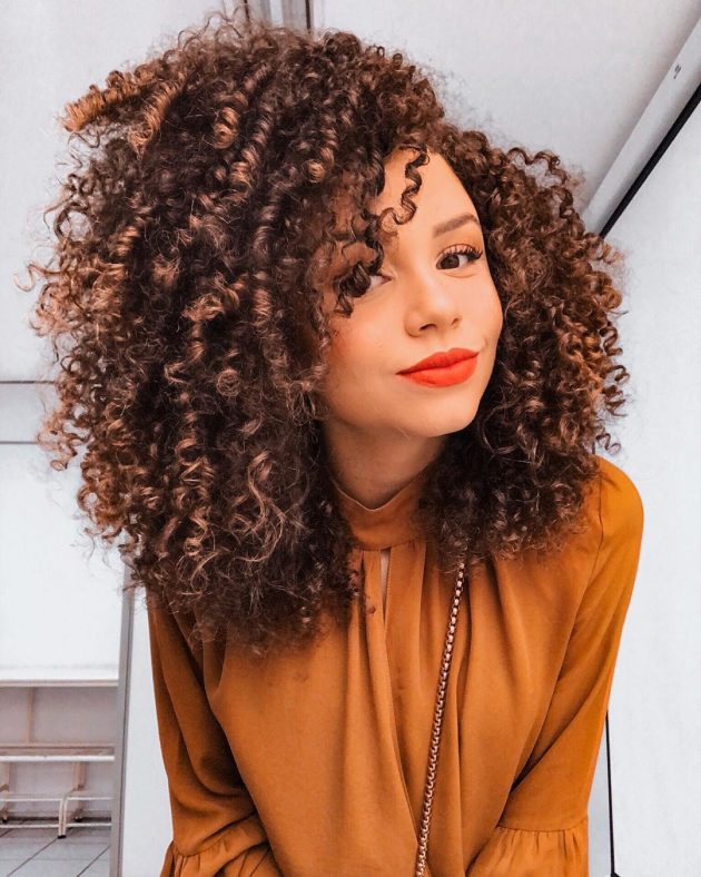 cabelo desfiado Jovem com expressão sorridente posando com blusa marrom e usando batom vermelho.