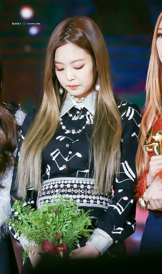 Foto da cantora Jennie do BLACKPINK. Ela usa um vestido preto com detalhes brancos e cabelo solto repartido em zigue-zague. Ela olha para baixo e não sorri para a foto.