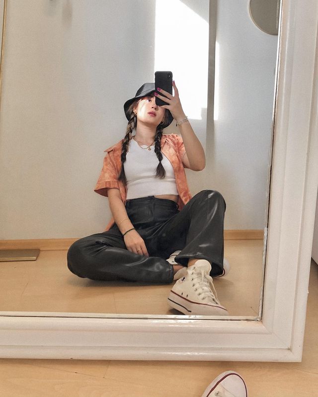 Selfie no espelho de uma mulher sentada no chão. Ela usa uma camiseta branca, camisa laranja tie dye como sobreposição, calça de couro preta, tênis branco de cano alto e bucket hat. Ela olha para o celular e não sorri.