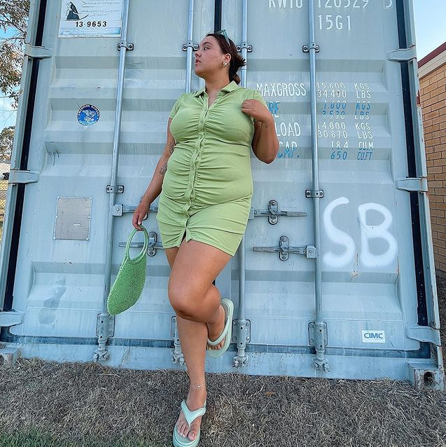 Garota usando vestido verde de botões frontais com chinelo de plataforma segurando uma bolsa verde. Ela está olhando para o lado, em pé com uma das pernas dobradas.