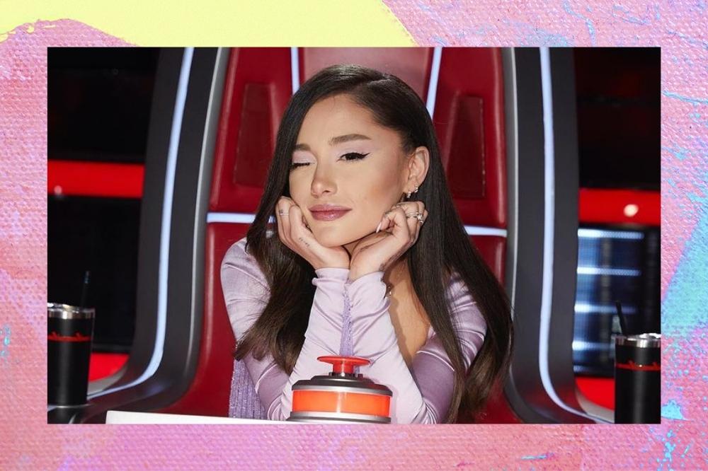 Montagem com a foto da Ariana Grande com o fundo rosa e amarelo. Ela usa um vestido lilás, cabelo solto e maquiagem com delineado preto e gloss. Ela está sentada na cadeira do The Voice com o rosto apoiado nas mãos, olha para a câmera e pisca com um olho.