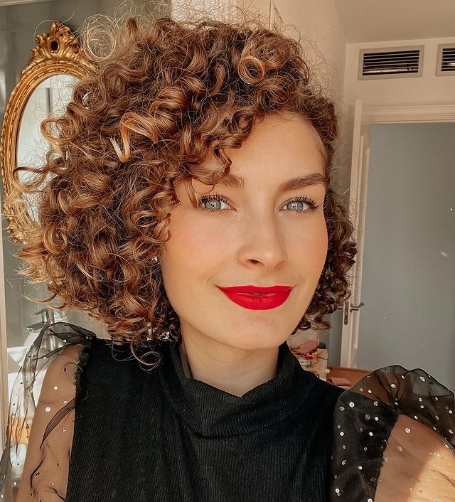 cabelo cacheado iluminado cobre Selfie de uma mulher. Ela usa uma blusa preta com manga bufante de tule, maquiagem básica com batom vermelho e cabelo cacheado curtinho iluminado em tom de cobre. Ela olha para a câmera e sorri levemente.