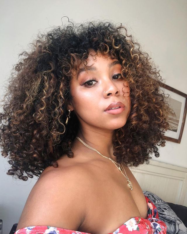 cabelo cacheado iluminado Selfie de uma mulher. Ela usa uma blusa vermelha com flores, colares prata, maquiagem natural e cabelo cacheado solto iluminado. Ela olha para a câmera e não sorri.