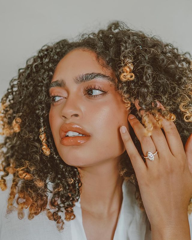 cabelo cobre iluminado Selfie de uma mulher. Ela usa uma camisa branca, maquiagem simples com gloss e cabelo cacheado solto iluminado em tom de cobre. Ela está com a mão esquerda no rosto com anel no dedo anelar, olha para o lado e não sorri.