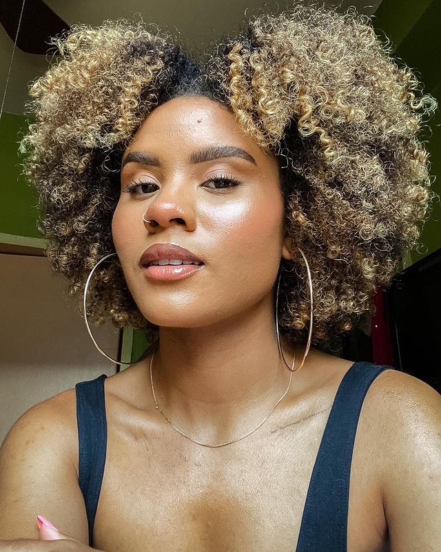 cabelo-crespo-iluminado Selfie de uma mulher. Ela usa uma blusa de alça preta, brinco de argola, maquiagem natural e cabelo crespo solto iluminado. Ela olha para a câmera e não sorri.