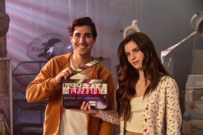 henry zaga giulia be depois do universo netflix Henry Zaga e Giulia Be nos bastidores de Depois do Universo; os dois estão seguando uma claquete com o nome do filme; Henry sorri e usa uma blusa branca com casaco laranja por cima enquanto Giulia usa uma blusa branca com casaco branco com detalhes de flores vermelhas e sorri levemente