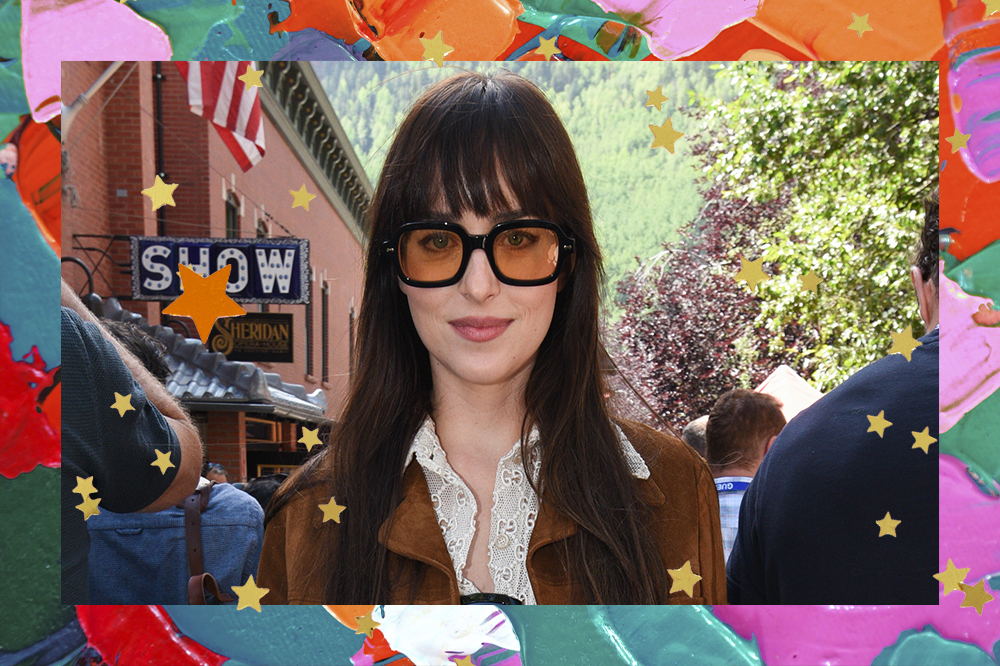 Dakota Johnson usando óculos de lentes alaranjadas, com expressão facial séria, camisa branca e jaqueta marrom. O fundo da montagem possui tintas coloridas e estrelinhas douradas