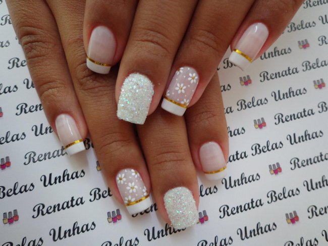 Foto de unhas decoradas com esmalte branco, decoração de flores no dedo anelar e fitilhos dourados.